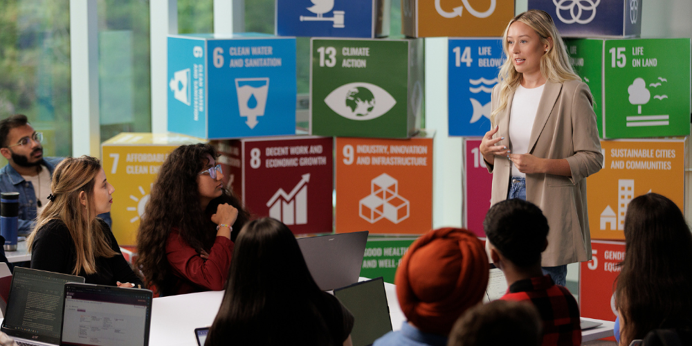 A student presents standing in front of colourful boxes depicting sustainability goals