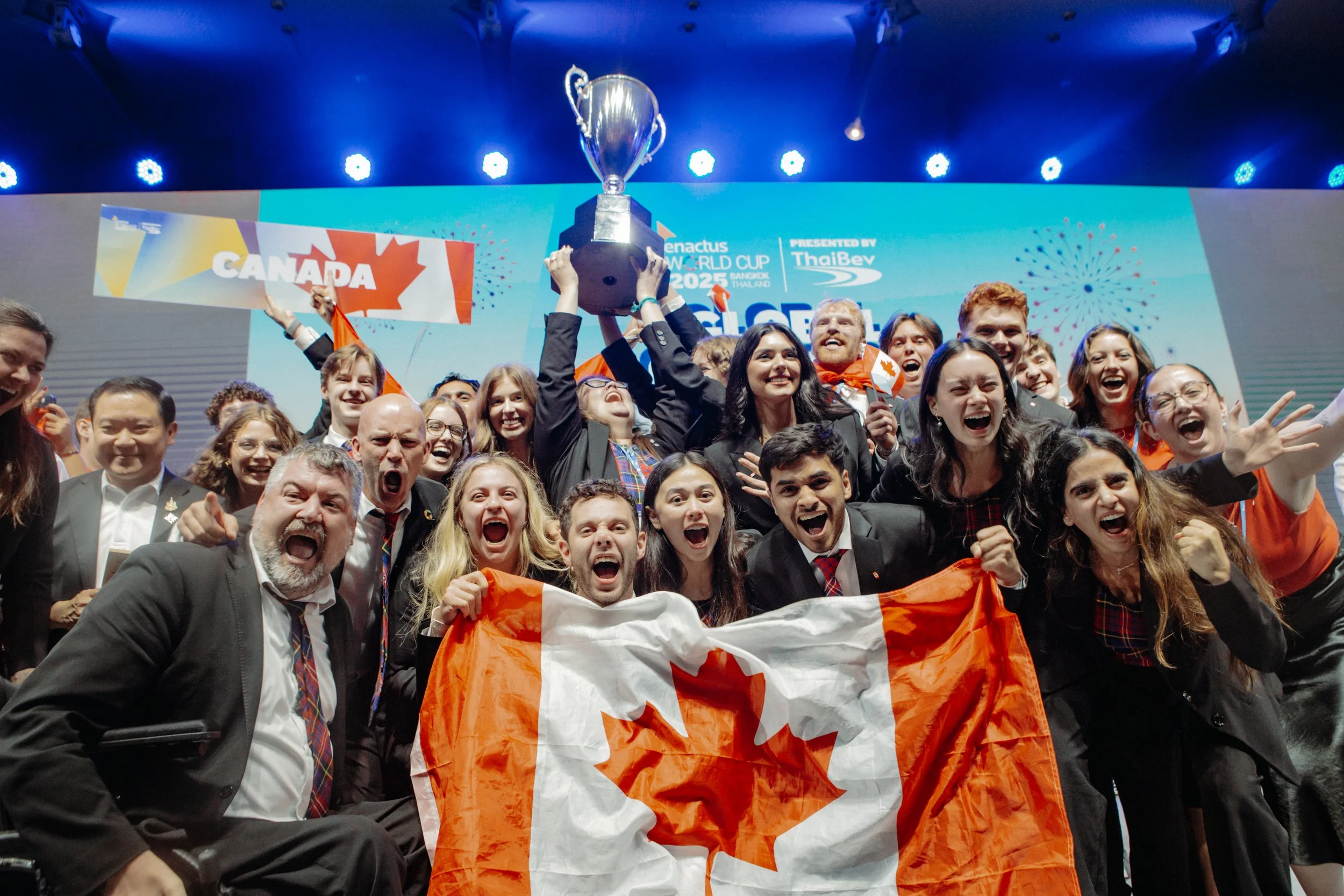 A large group is smiling and cheering. One person holds a trophy aloft while others hold the Canadian flag.