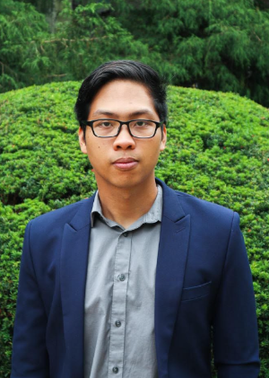 A student posing for a picture, with a large green bush in the background